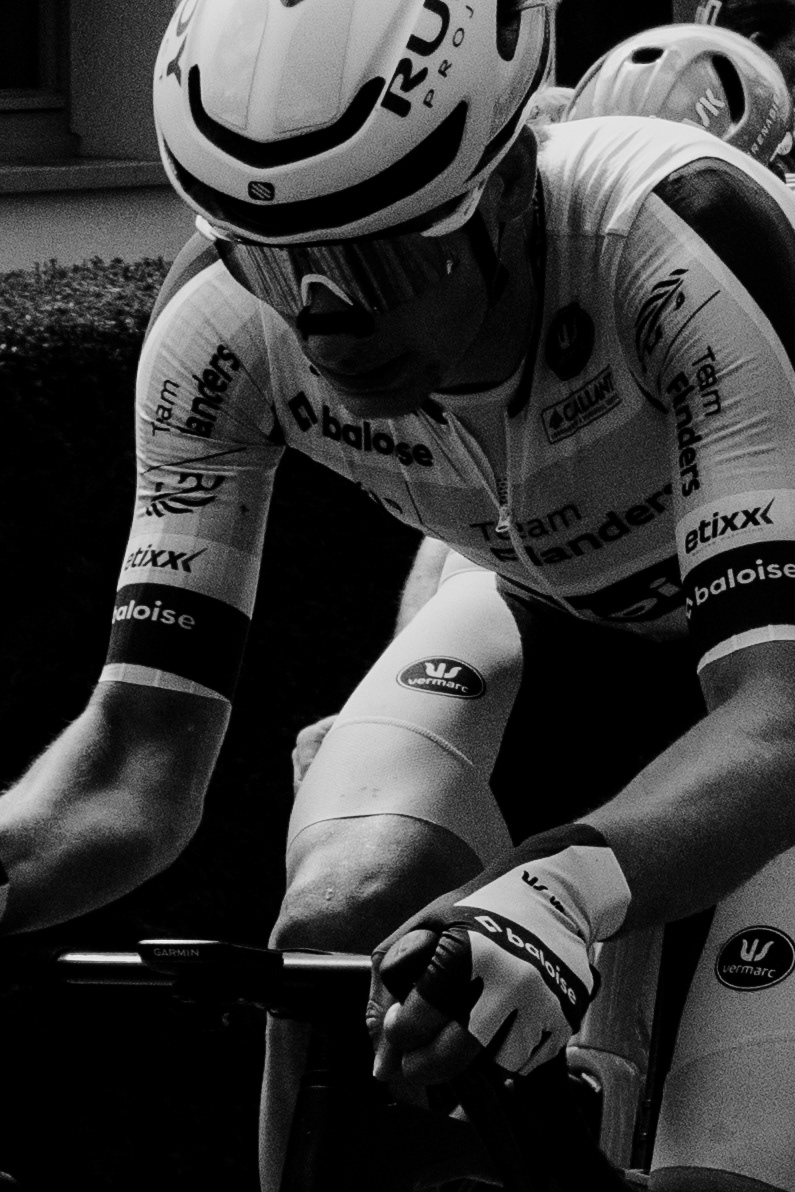 Black and white close-up of a cyclist in a Team Flanders–Baloise jersey, gripping the handlebars
